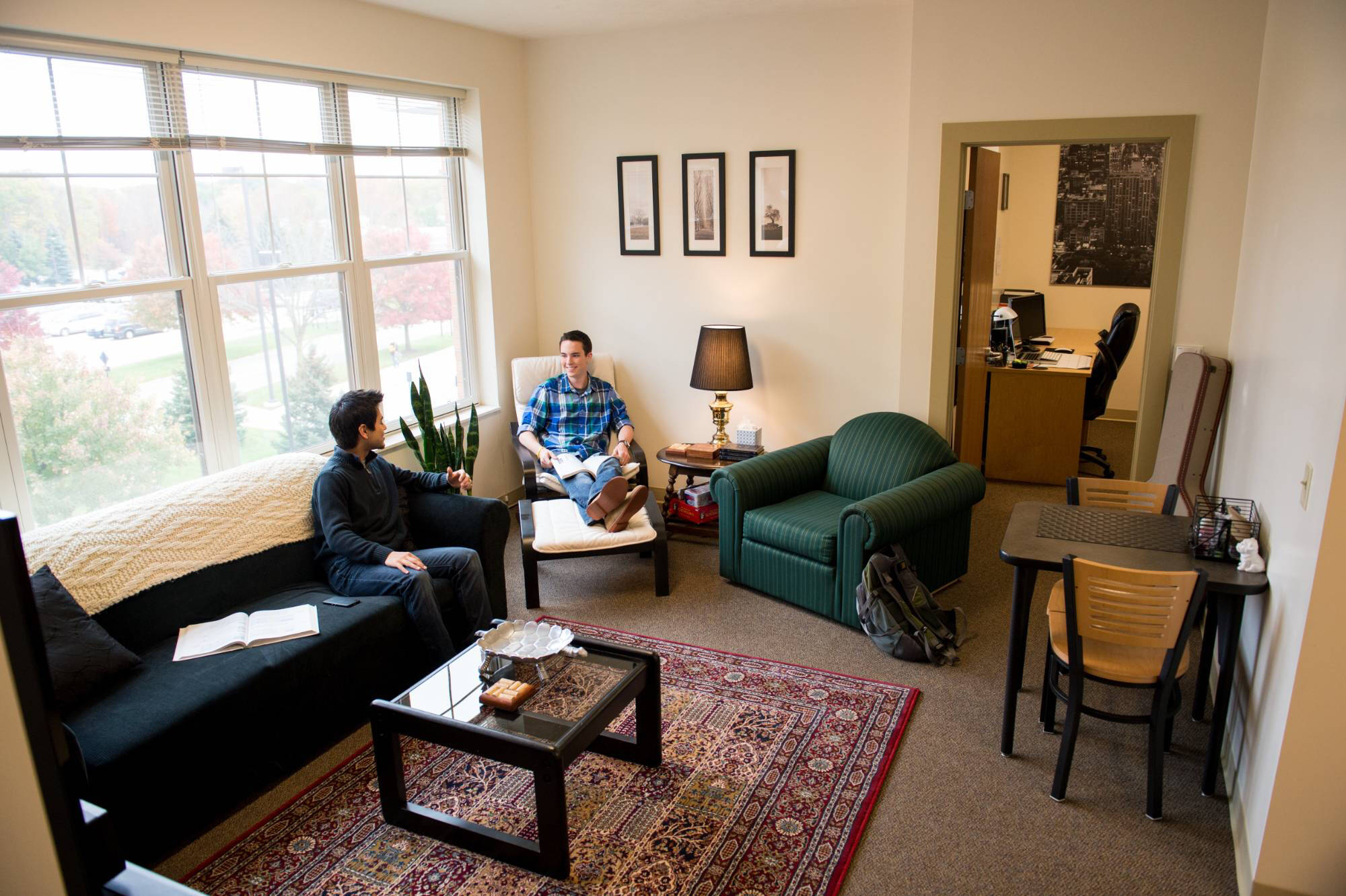Two people converse in a cozy, sunlit apartment living room with modern decor. A sofa, armchairs, and a coffee table on a patterned rug create a warm atmosphere.